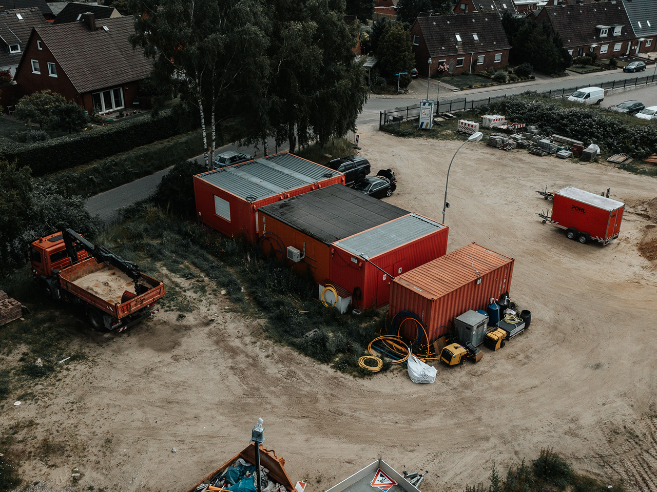 Standort Heide: Vorne im Bild liegen mehrere orangefarbene Container mit Geräten und Schläuchen auf einem Baugrundstück. In der Mitte parkt ein roter Lkw; rechts daneben stehen Autos an einer Straße. Im Hintergrund stehen Einfamilienhäuser mit Gärten.