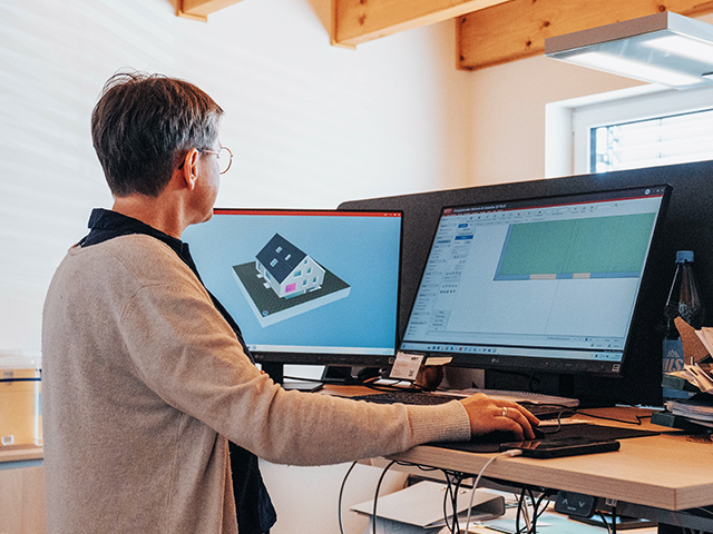 Eine Person steht an einem höhenverstellbaren Schreibtisch und arbeitet an zwei Monitoren. Auf dem linken Bildschirm ist ein 3D-Modell eines Hauses zu sehen, auf dem rechten ein technisches Diagramm. Der Raum ist hell und modern eingerichtet.