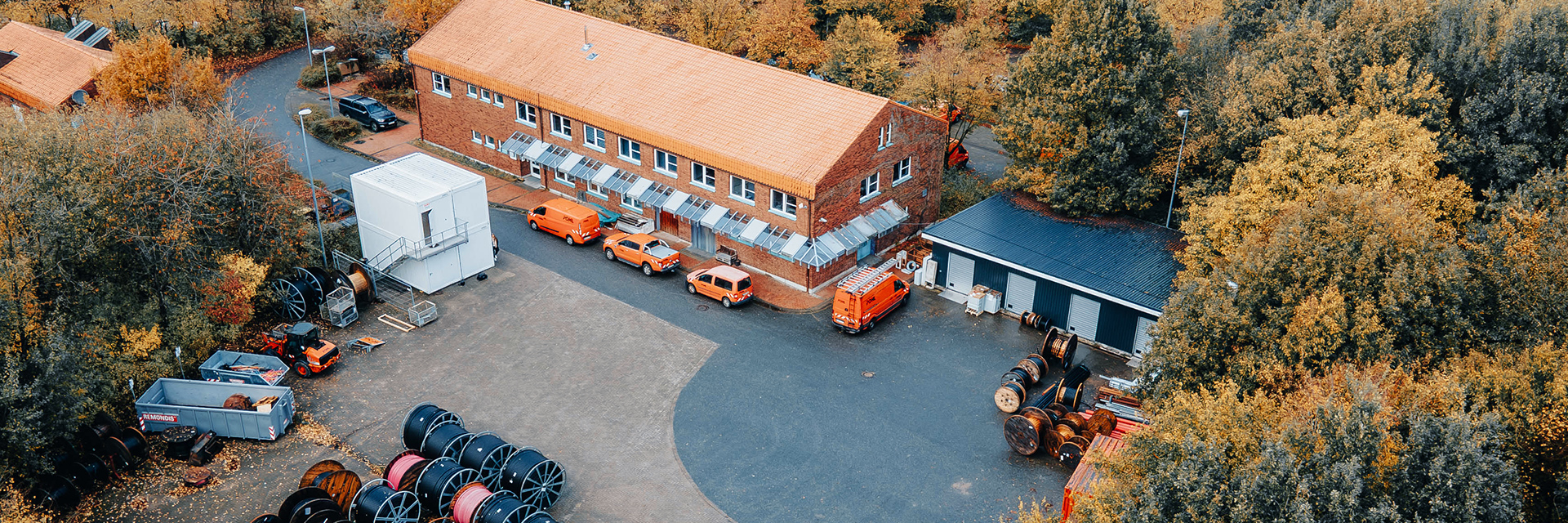Standort Schleswig: Eine Luftaufnahme eines Firmengeländes mit einem langen Backsteingebäude, mehreren orangefarbenen Firmenfahrzeugen und vielen großen Kabeltrommeln auf dem Hof. Rundherum stehen herbstlich gefärbte Bäume.