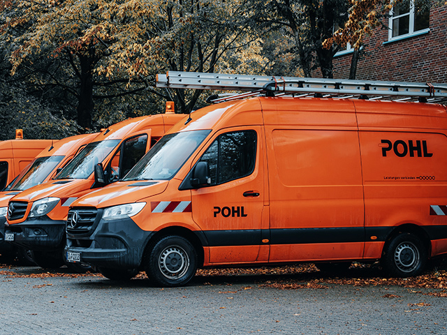 Standort Schleswig: Drei orangefarbene Transporter der Firma POHL stehen nebeneinander auf einem Parkplatz. Jeder Wagen trägt das Firmenlogo und auf dem Dach eine Leiter. Im Hintergrund stehen herbstliche Bäume mit gelben Blättern.