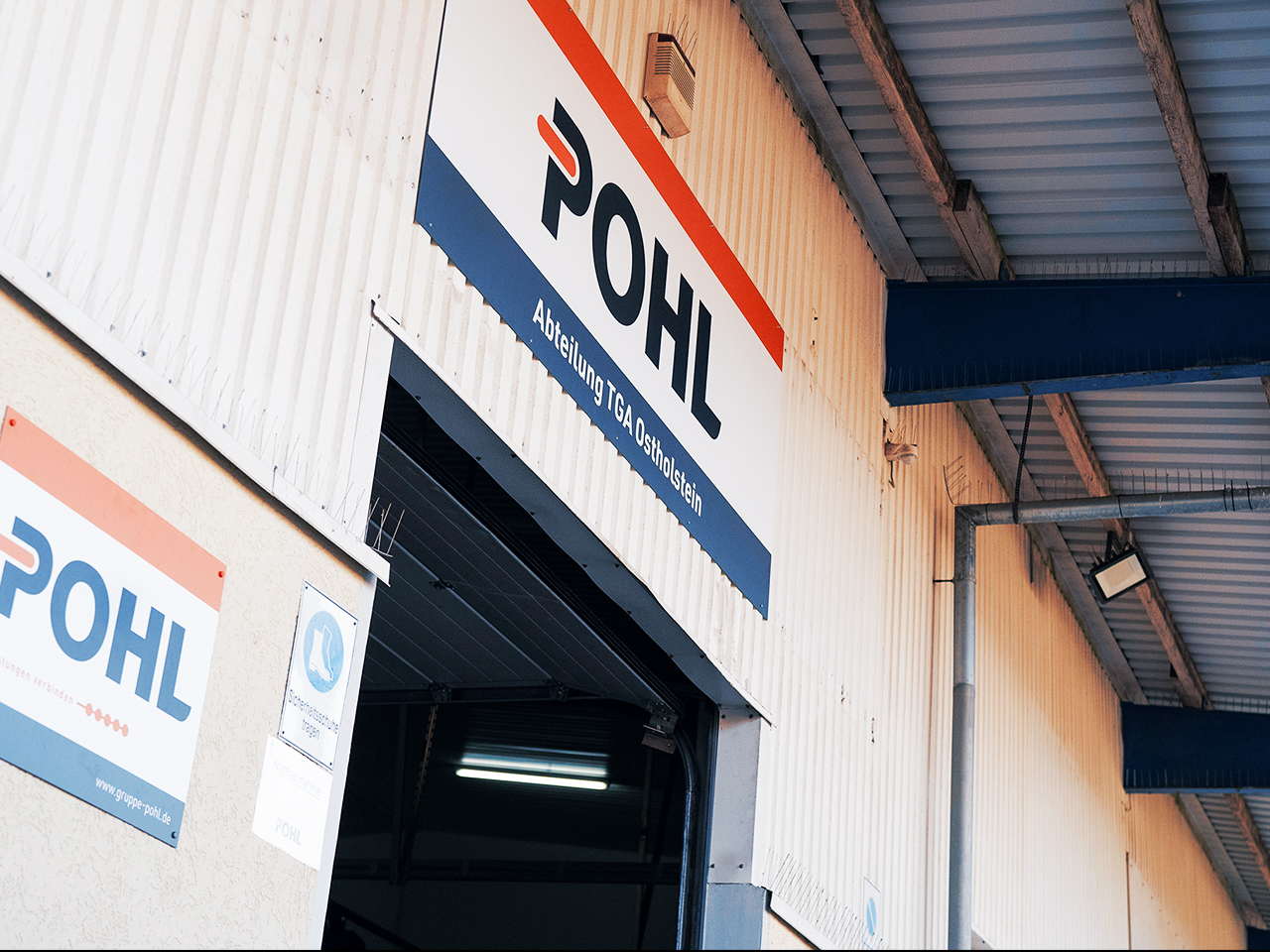 Standort Ahrensbök: Oben rechts über einem Hallentor hängt ein großes POHL-Schild. Links daneben sind weitere POHL-Schilder an der Wand; oben verläuft das Metall­dach.