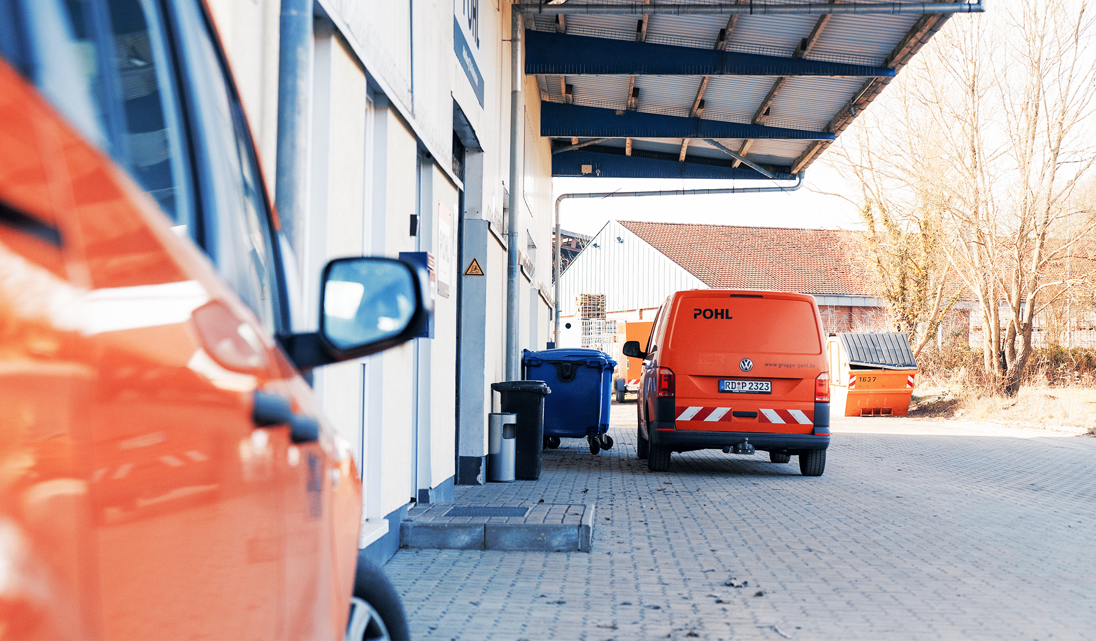 Standort Ahrensbök: In der Mitte unter einem Vordach steht hinten ein orangefarbener POHL-Transporter. Links am Rand ist die Seite eines weiteren orangefarbenen POHL-Fahrzeugs zu sehen; rechts im Hintergrund stehen Müllbehälter und ein orangefarbener Container.