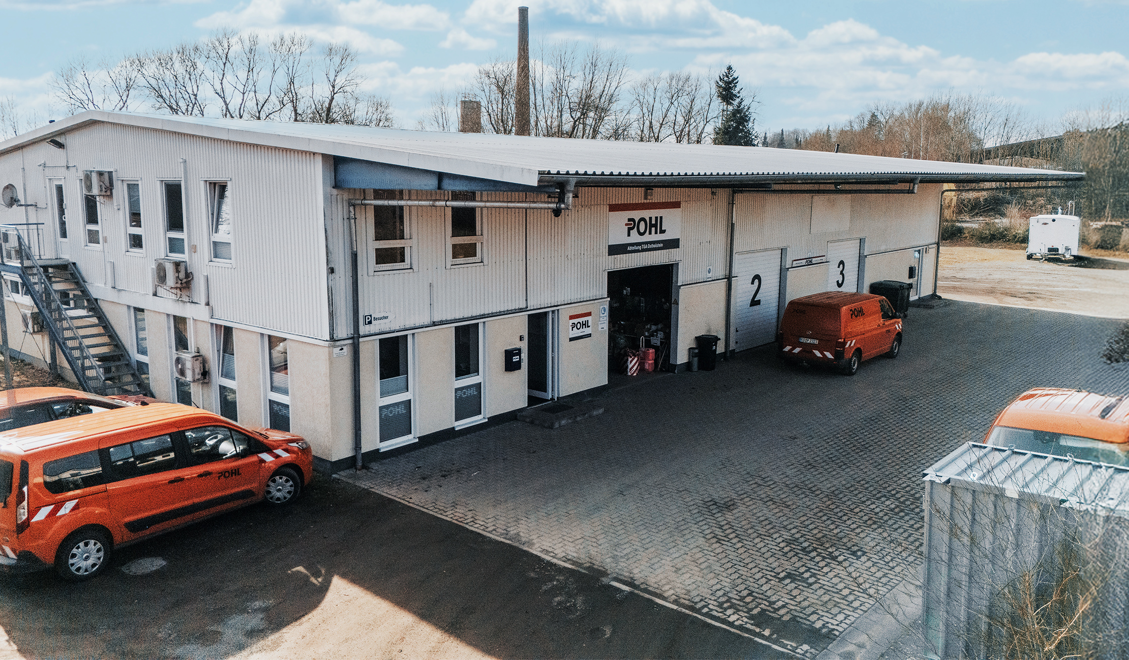 Standort Ahrensbök: In der Mitte steht ein zweigeschossiges Gebäude mit seitlicher Treppe und nummerierten Toren; vor den Toren parken mehrere orange POHL-Fahrzeuge. Rechts am Rand öffnet sich ein Hof; im Hintergrund stehen Bäume.