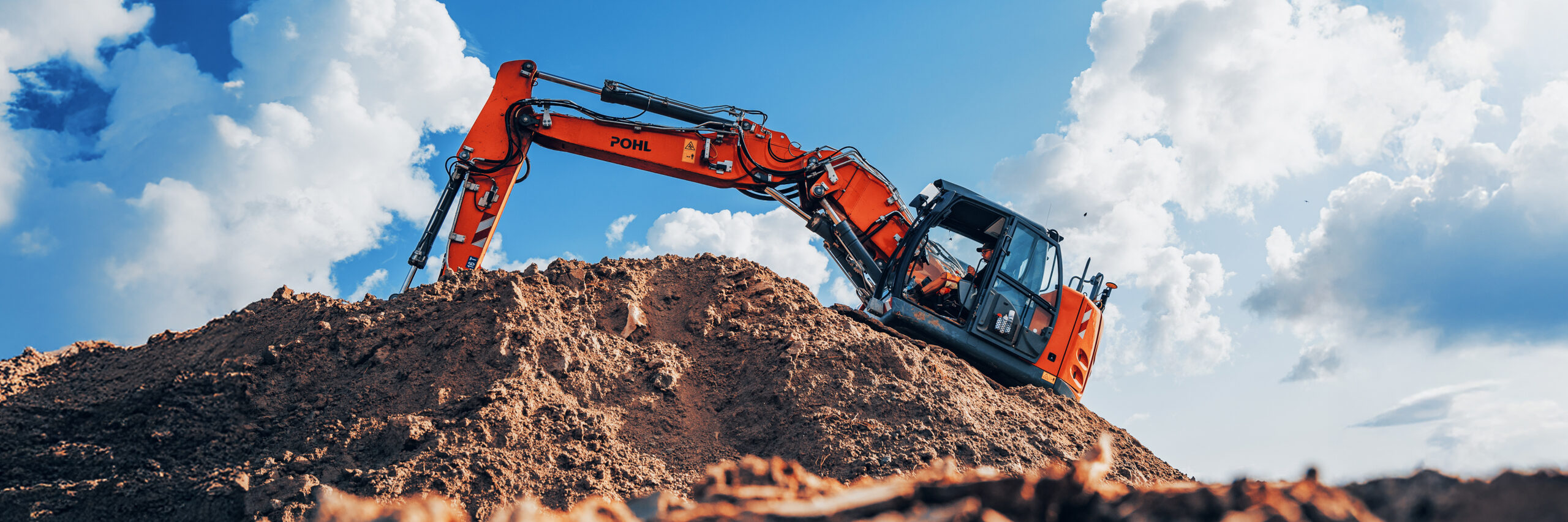 Ein großer orangefarbener Bagger von POHL arbeitet auf einem Erdhügel. Der Arm der Maschine ragt in den Himmel, im Hintergrund sind helle Wolken zu sehen.