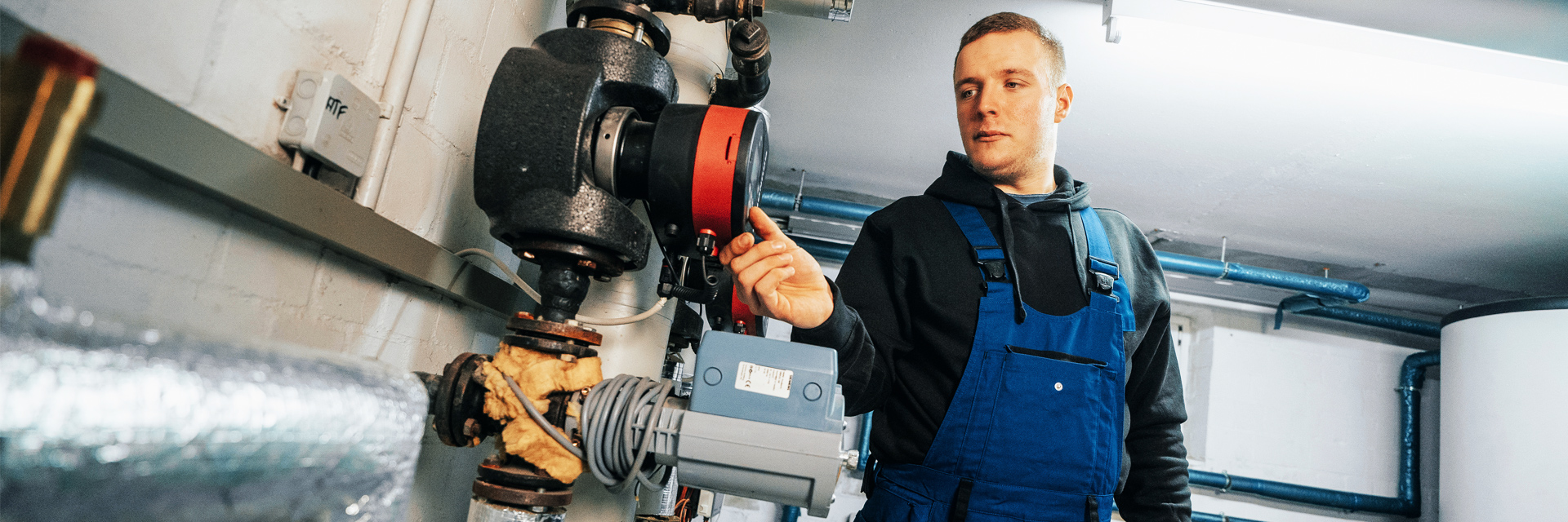In der Mitte zeigt ein Mann in blauer Latzhose und dunklem Hoodie auf ein rotes rundes Teil an einer Rohrleitung. Links im Bild verlaufen isolierte Rohre an einer weißen Wand. Rechts im Bild stehen blaue Leitungen und ein großer weißer Behälter. Im Hintergrund ziehen sich weitere Leitungen unter der Decke entlang. Vorne im Bild ist ein silbrig umwickeltes Rohr angeschnitten.