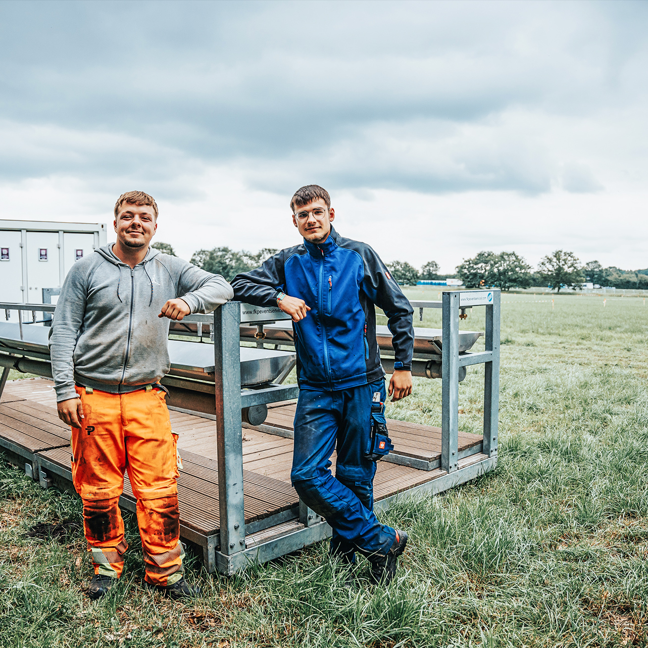 Auf einer Wiese lehnen zwei POHL-Kollegen an einem modularen Bühnensteg; links orange Arbeitshose, rechts blaue Arbeitskleidung; grauer Himmel.