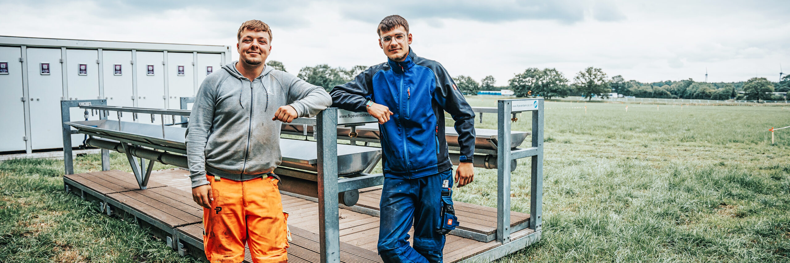 Auf einer Wiese lehnen zwei POHL-Kollegen an einem modularen Bühnensteg; links orange Arbeitshose, rechts blaue Arbeitskleidung; grauer Himmel.