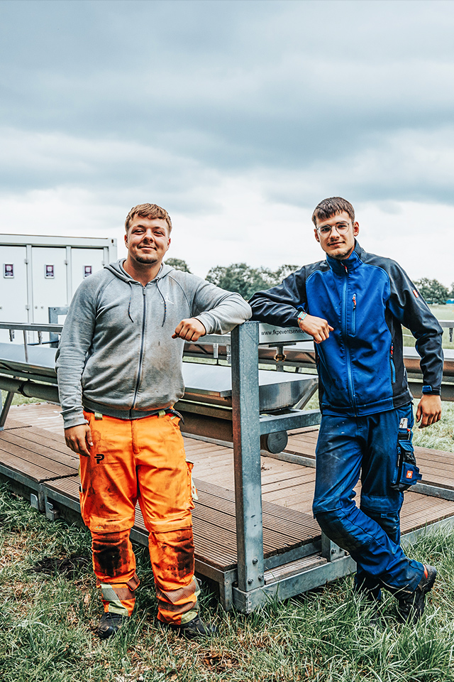 Auf einer Wiese lehnen zwei POHL-Kollegen an einem modularen Bühnensteg; links orange Arbeitshose, rechts blaue Arbeitskleidung; grauer Himmel.
