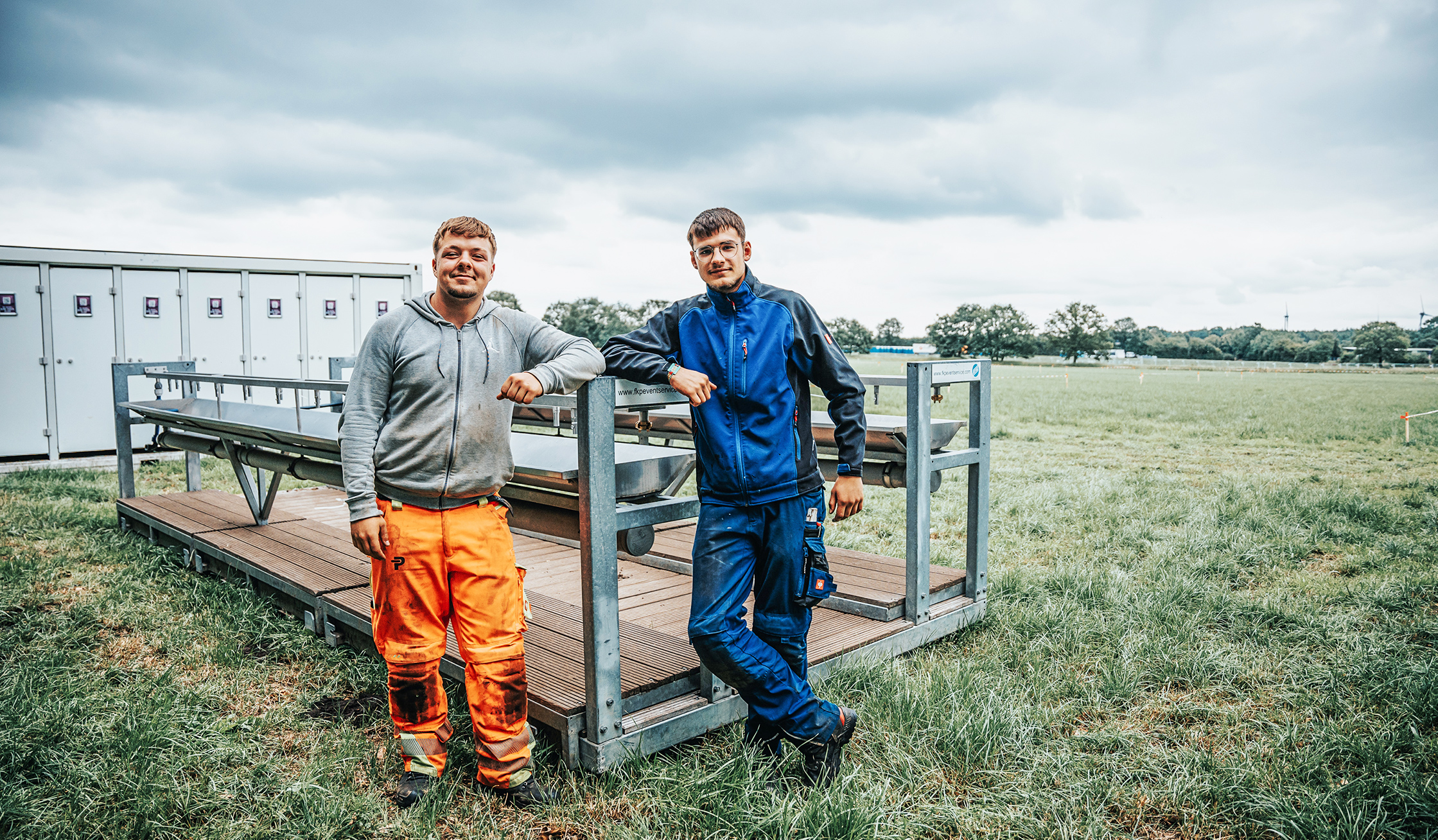 Auf einer Wiese lehnen zwei POHL-Kollegen an einem modularen Bühnensteg; links orange Arbeitshose, rechts blaue Arbeitskleidung; grauer Himmel.