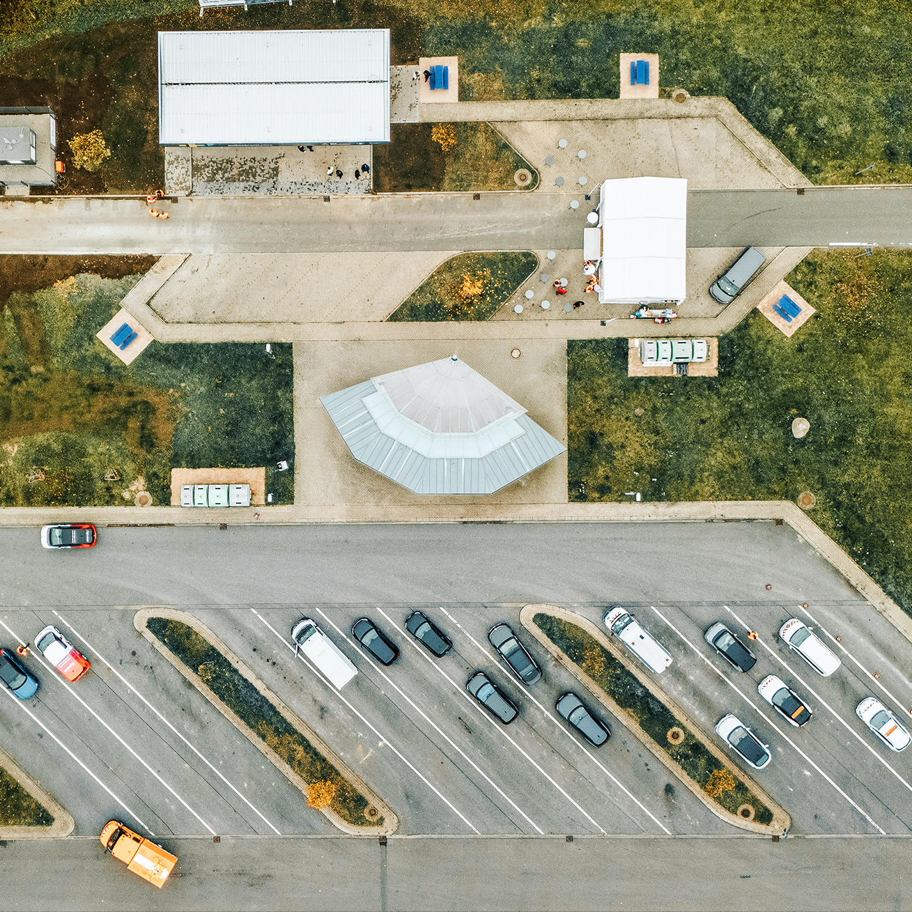 Referenz Autostrom Depenauer Moor: Luftbild eines Parkplatzes: In der Mitte stehen Autos in schrägen Parkreihen. Oben und unten verlaufen Zufahrtsstraßen; am oberen Rand fahren zwei Fahrzeuge, darunter ein orangefarbenes POHL-Fahrzeug. Links und rechts liegen Grünflächen mit einzelnen Bäumen.