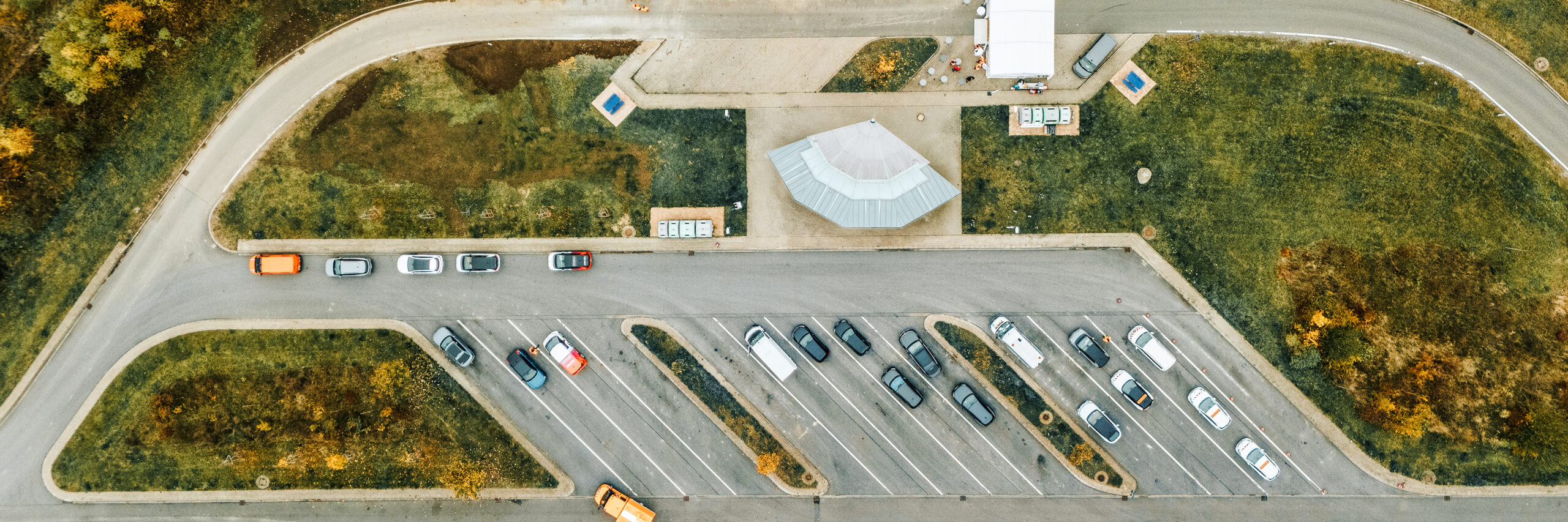 Referenz Autostrom Depenauer Moor: Luftbild eines Parkplatzes: In der Mitte stehen Autos in schrägen Parkreihen. Oben und unten verlaufen Zufahrtsstraßen; am oberen Rand fahren zwei Fahrzeuge, darunter ein orangefarbenes POHL-Fahrzeug. Links und rechts liegen Grünflächen mit einzelnen Bäumen.