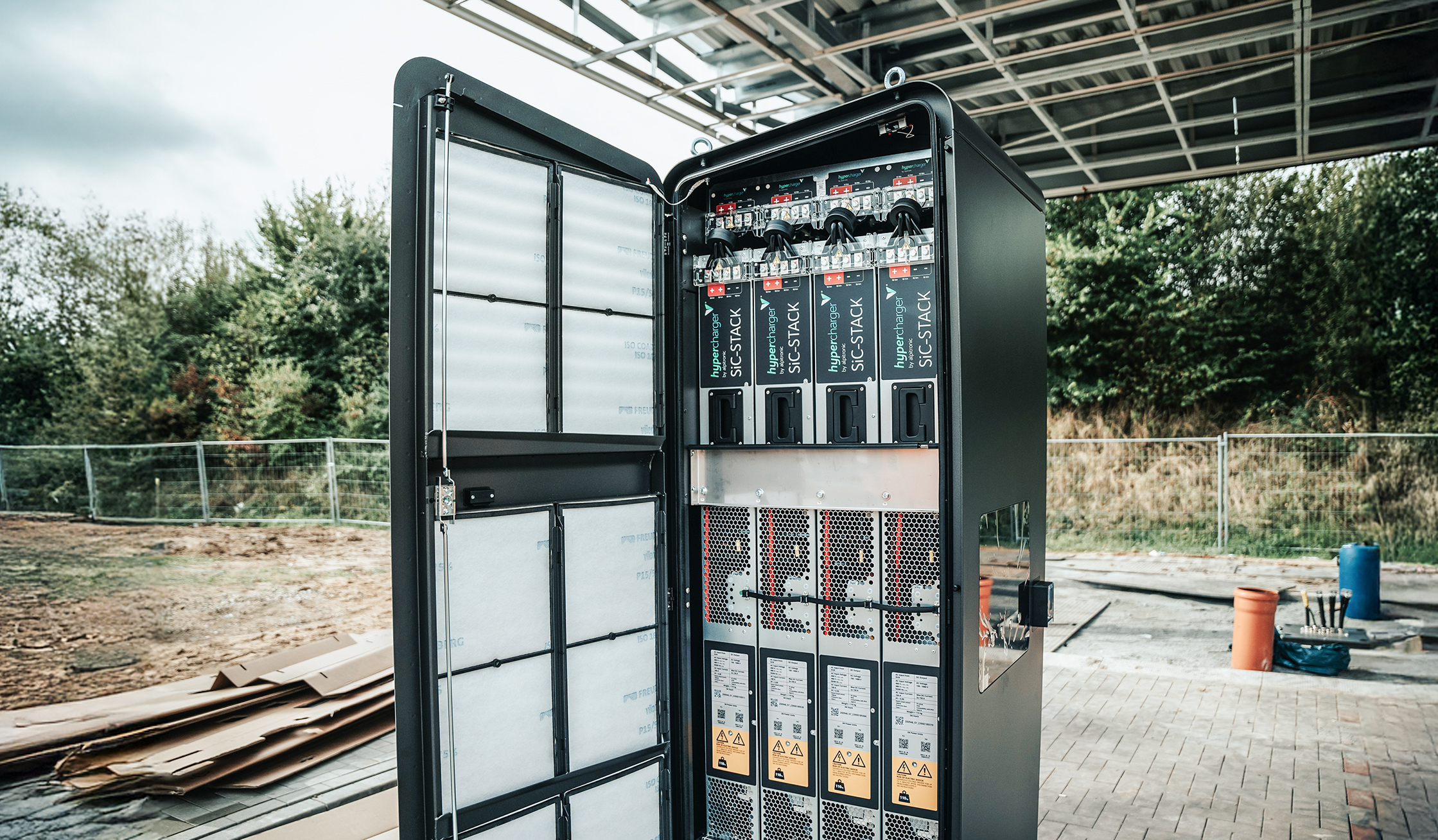 Referenz Autostrom Depenauer Moor: Vorne links ist ein geöffneter, hoher Technikschrank mit vielen Einschüben, Kabeln und Lüftungsgittern zu sehen. Rechts daneben verläuft eine Baufläche mit Kartonstapeln; im Hintergrund stehen Zäune und Büsche.