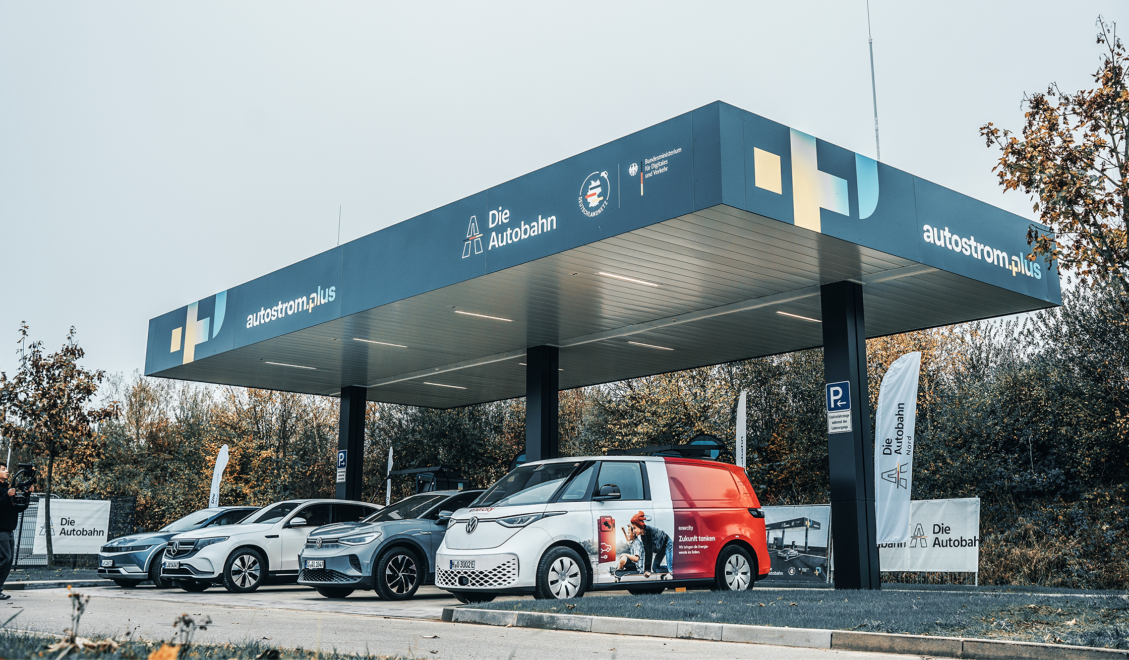 Referenz Autostrom Depenauer Moor: In der Mitte steht ein dunkles Überdach mit dem Schriftzug „autostrom.plus“. Darunter parken nebeneinander mehrere Autos; links und rechts stehen Fahnen mit „Die Autobahn“. Hinten bildet eine Baumreihe den Hintergrund.