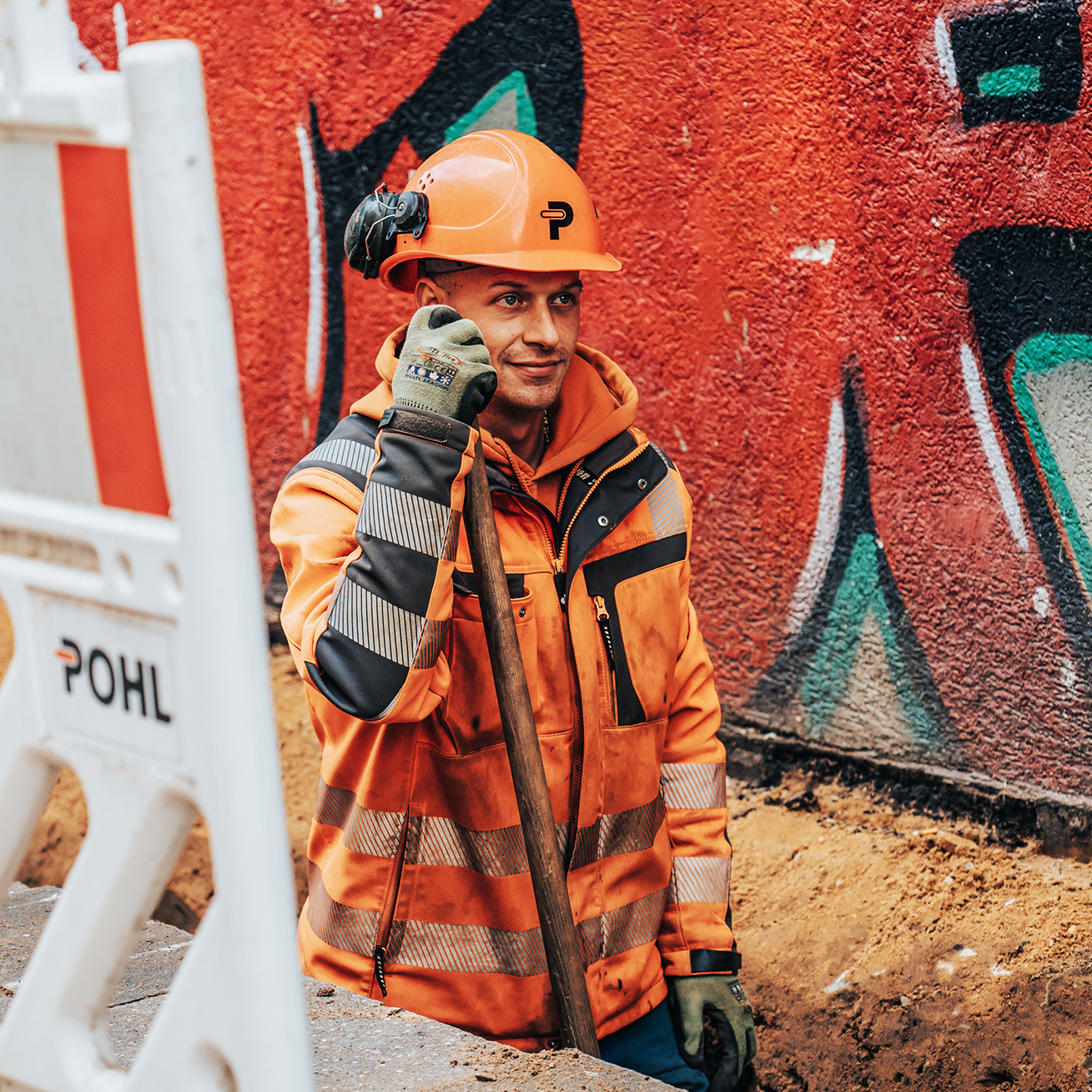 In einem Rohrgraben lehnt ein POHL-Mitarbeiter mit Helm an einer Schaufel; links eine POHL-Absperrbake, rechts eine rote Graffitiwand.