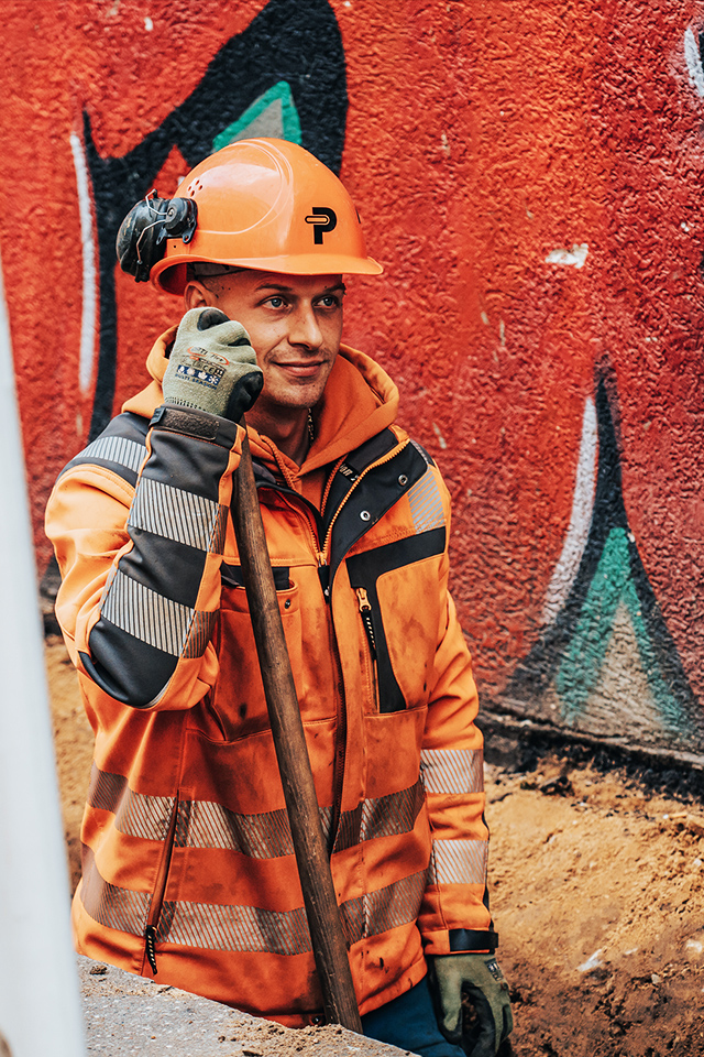 In einem Rohrgraben lehnt ein POHL-Mitarbeiter mit Helm an einer Schaufel; links eine POHL-Absperrbake, rechts eine rote Graffitiwand.