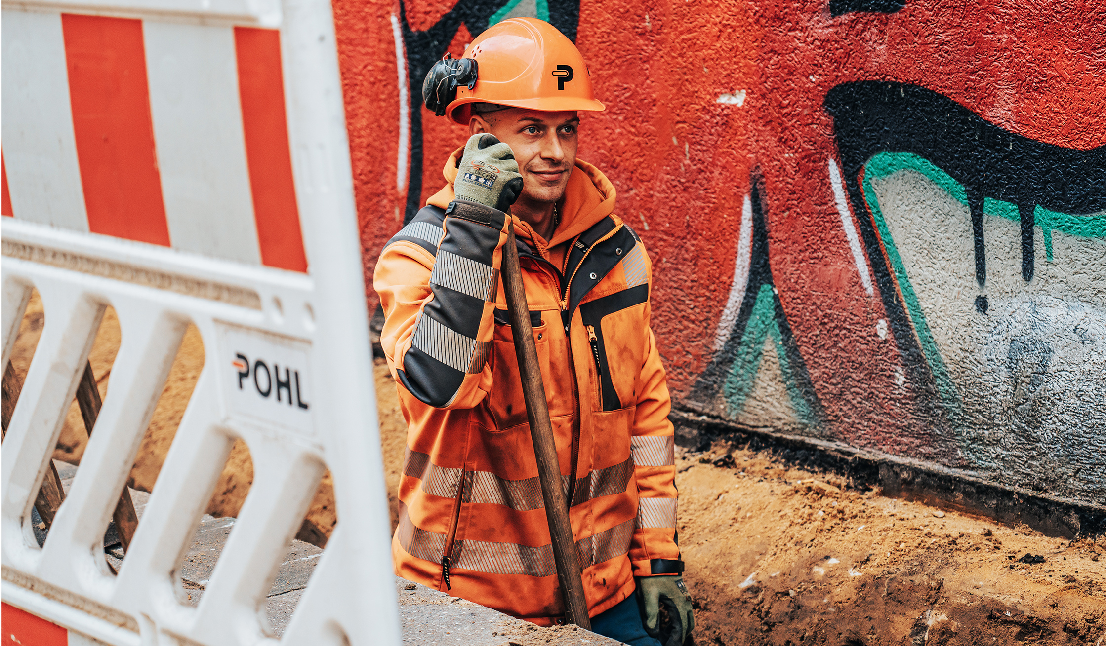 In einem Rohrgraben lehnt ein POHL-Mitarbeiter mit Helm an einer Schaufel; links eine POHL-Absperrbake, rechts eine rote Graffitiwand.