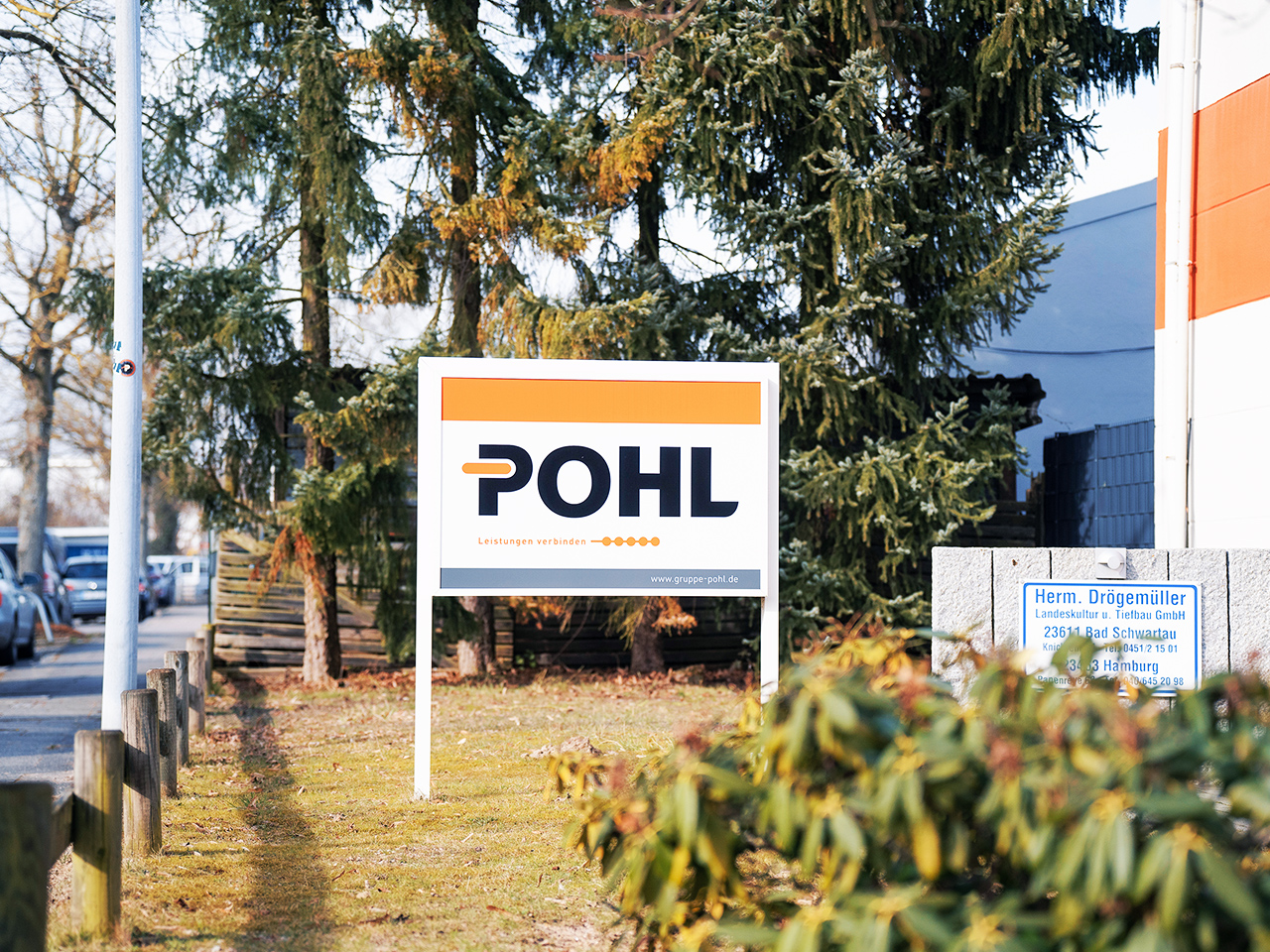 Standort Bad Schwartau: In der Mitte steht auf einer Rasenfläche ein weißes Schild mit POHL-Logo. Links am Rand verläuft eine Straße mit geparkten Autos; im Hintergrund stehen Bäume.