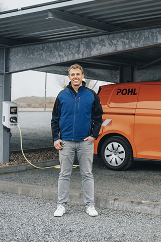 In der Mitte rechts steht ein orangefarbenes POHL-Fahrzeug mit sichtbarem POHL-Schriftzug unter einem überdachten Stellplatz. Links im Bild steht ein Mann in blauer Jacke vor einer grauen Stütze. An der Stütze ist eine weiße Ladesäule mit Kabel montiert. Rechts im Bild parken weitere Fahrzeuge. Vorne im Bild liegt das Kabel locker auf dem Kiesboden.