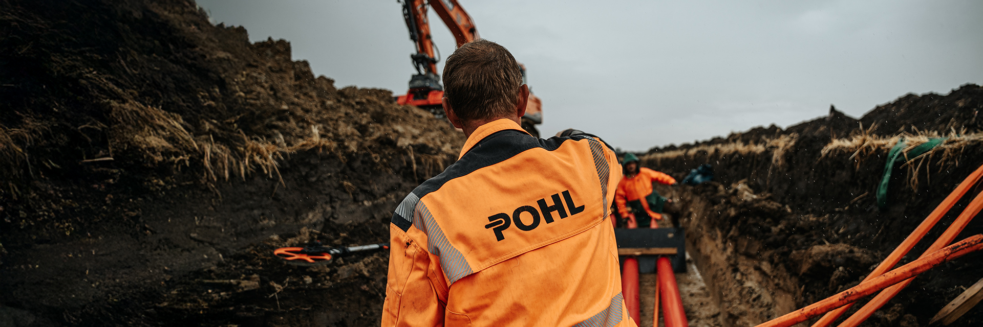 Von hinten ist in der Mitte ein POHL-Mitarbeiter mit orangefarbener Jacke zu sehen; vorne rechts im Bild liegen orangefarbene Rohre, links oben arbeitet ein orangefarbener POHL-Bagger an einer Erdwand.