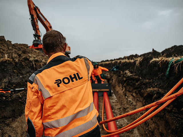 Von hinten ist in der Mitte ein POHL-Mitarbeiter mit orangefarbener Jacke zu sehen; vorne rechts im Bild liegen orangefarbene Rohre, links oben arbeitet ein orangefarbener POHL-Bagger an einer Erdwand.