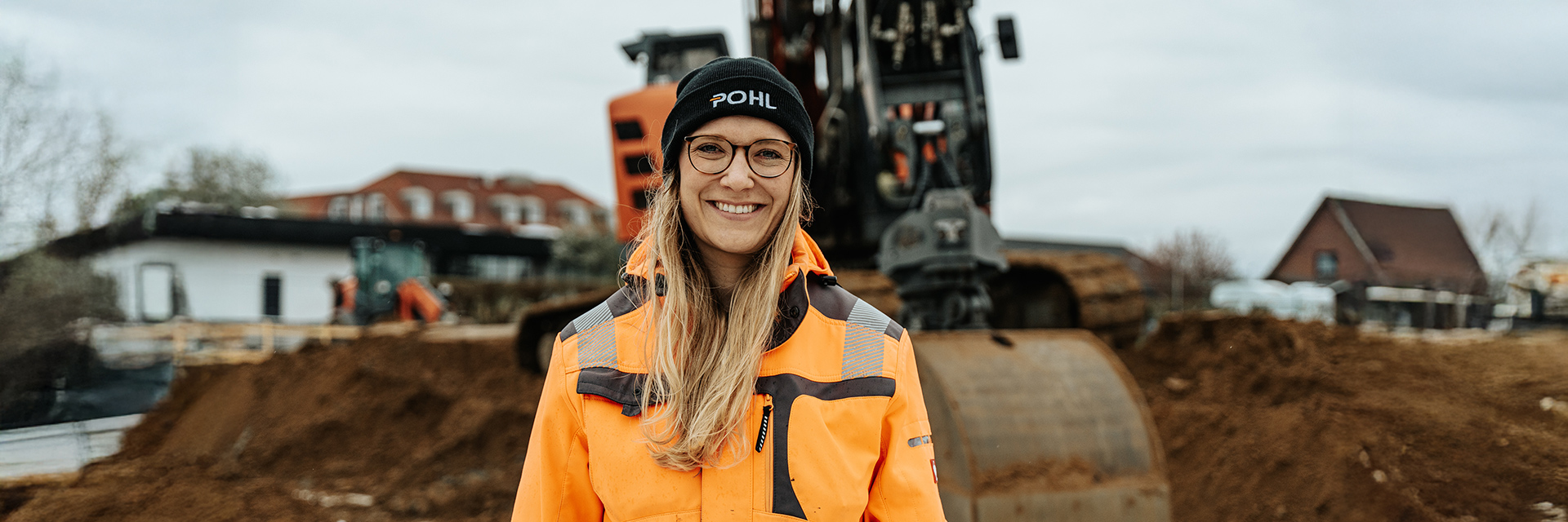 In der Mitte steht eine Frau mit schwarzer POHL-Mütze und orangefarbener Jacke; hinter ihr steht ein orangefarbener POHL-Bagger vor Erdhaufen und Gebäuden.