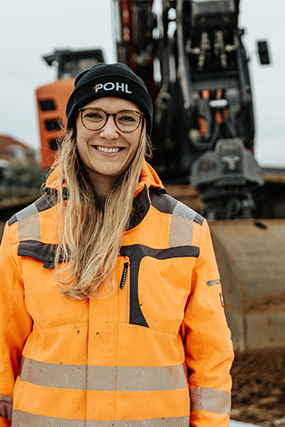 In der Mitte steht eine Frau mit schwarzer POHL-Mütze und orangefarbener Jacke; hinter ihr steht ein orangefarbener POHL-Bagger vor Erdhaufen und Gebäuden.