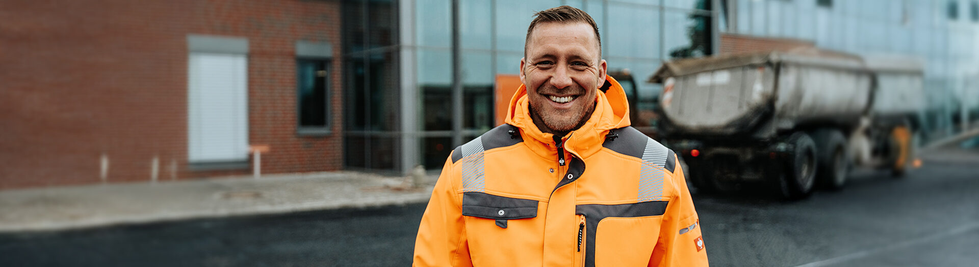 In der Mitte steht ein POHL-Mitarbeiter in orangefarbener Jacke vor einem Gebäude; rechts im Hintergrund parkt ein großer Anhänger auf frischem Asphalt, links sind Glasfassaden.