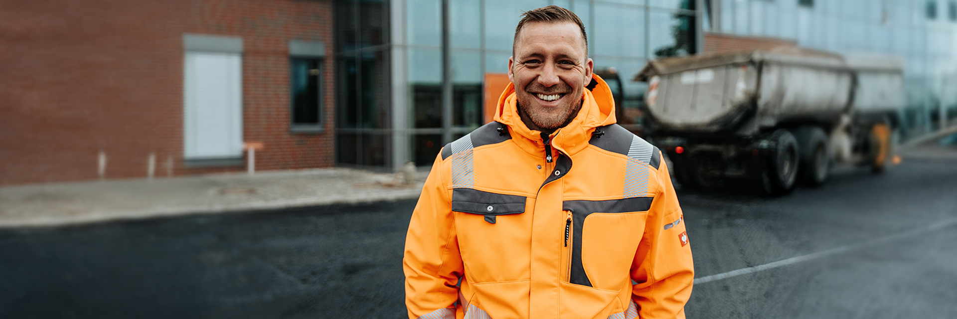 In der Mitte steht ein POHL-Mitarbeiter in orangefarbener Jacke vor einem Gebäude; rechts im Hintergrund parkt ein großer Anhänger auf frischem Asphalt, links sind Glasfassaden.