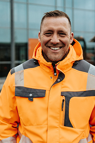 In der Mitte steht ein POHL-Mitarbeiter in orangefarbener Jacke vor einem Gebäude; rechts im Hintergrund parkt ein großer Anhänger auf frischem Asphalt, links sind Glasfassaden.