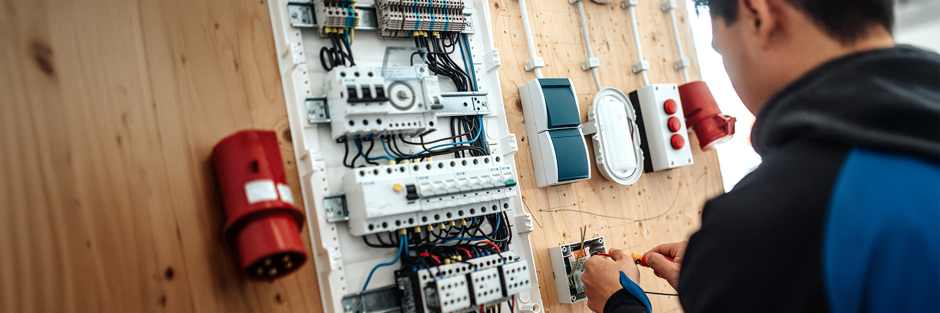 In der Mitte ist eine große, weiße Verteilerwand mit vielen Kabeln und Schaltern an einer Holzplatte zu sehen; rechts im Bild arbeitet eine Person mit einem Schraubendreher an einem offenen Kästchen.