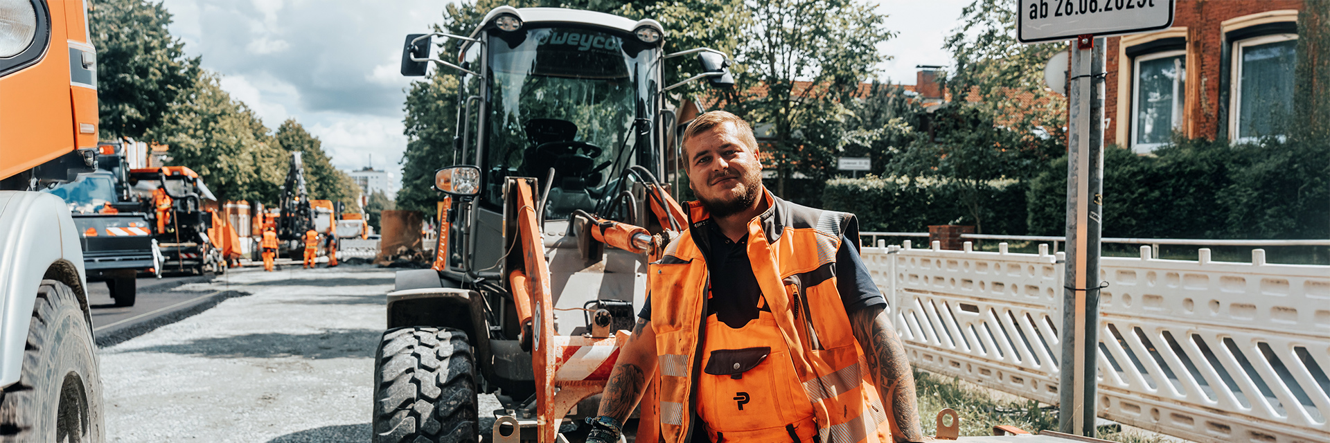 In der Mitte steht ein POHL-Mitarbeiter in orangefarbener Weste auf einer Straßenbaustelle neben einer Radladerschaufel. Links im Bild ragt das Vorderrad eines orangefarbenen Fahrzeugs ins Bild. Rechts im Bild steht ein Schildmast am Gehweg. Im Hintergrund arbeiten mehrere orangefarbene Maschinen und weiße Absperrelemente entlang der Straße.