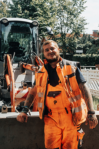 In der Mitte steht ein POHL-Mitarbeiter in orangefarbener Weste auf einer Straßenbaustelle neben einer Radladerschaufel. Links im Bild ragt das Vorderrad eines orangefarbenen Fahrzeugs ins Bild. Rechts im Bild steht ein Schildmast am Gehweg. Im Hintergrund arbeiten mehrere orangefarbene Maschinen und weiße Absperrelemente entlang der Straße.