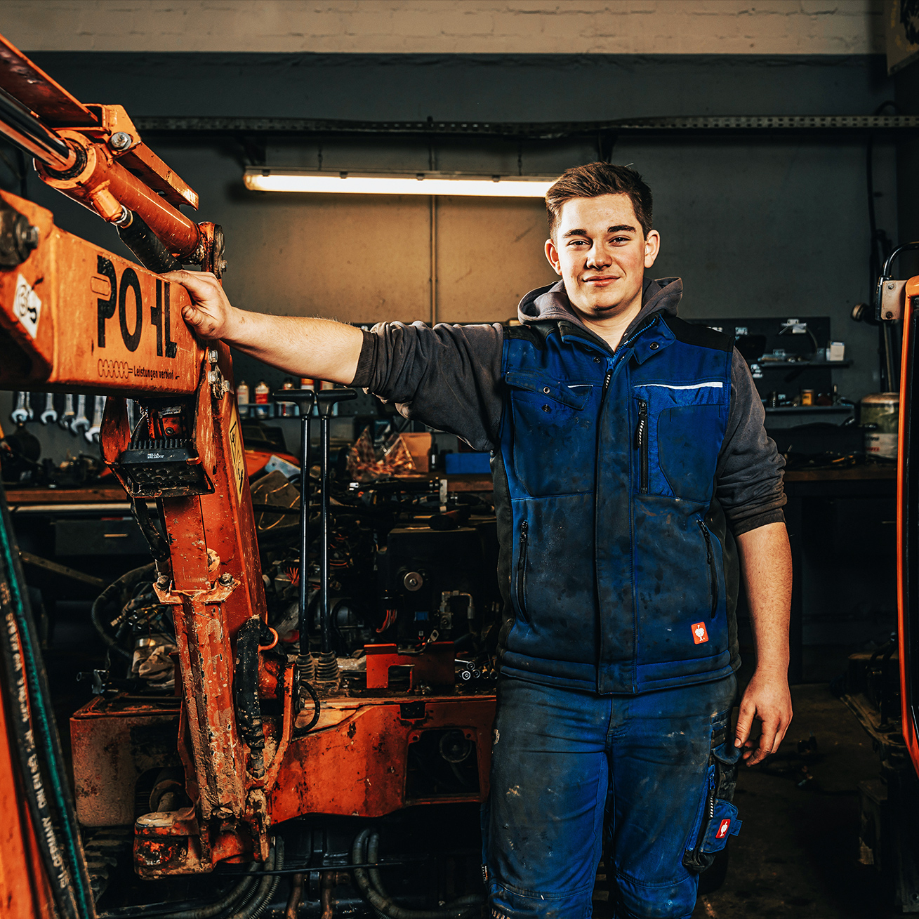 In der Werkstatt lehnt ein POHL-Mechatroniker an einem Baggerarm mit POHL-Schriftzug; um ihn herum Maschinen und Werkzeuge.