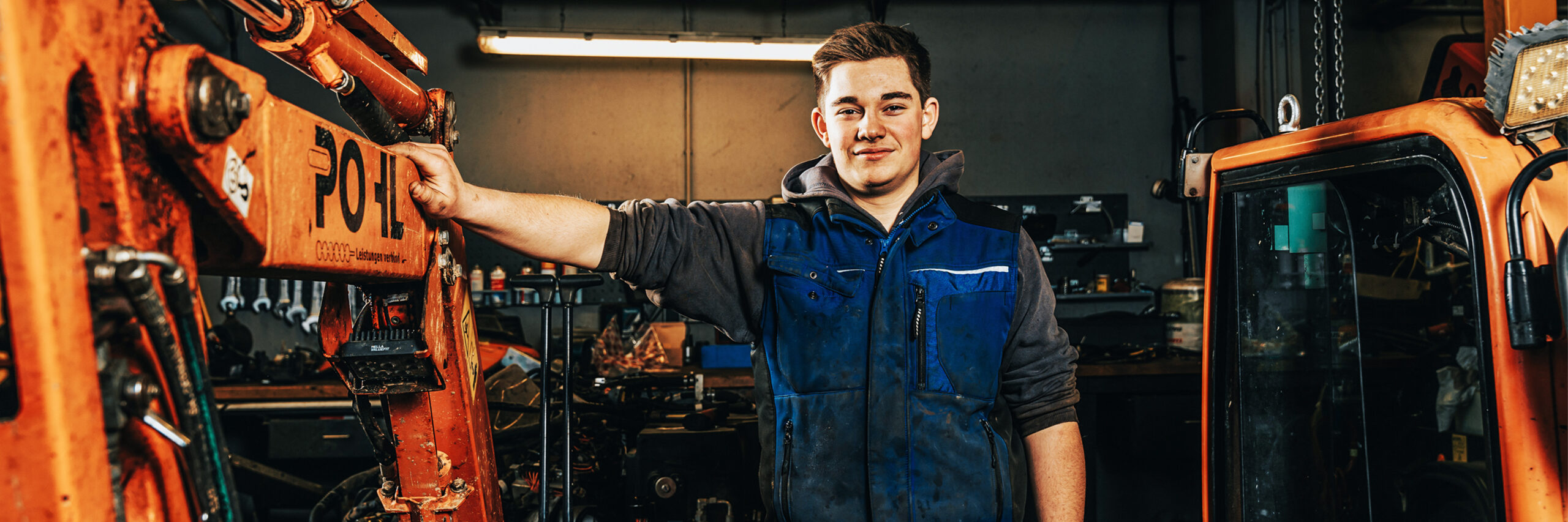 In der Werkstatt lehnt ein POHL-Mechatroniker an einem Baggerarm mit POHL-Schriftzug; um ihn herum Maschinen und Werkzeuge.