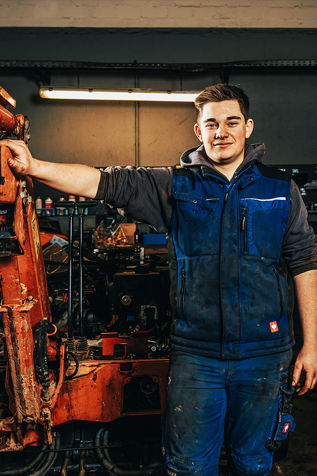 In der Werkstatt lehnt ein POHL-Mechatroniker an einem Baggerarm mit POHL-Schriftzug; um ihn herum Maschinen und Werkzeuge.