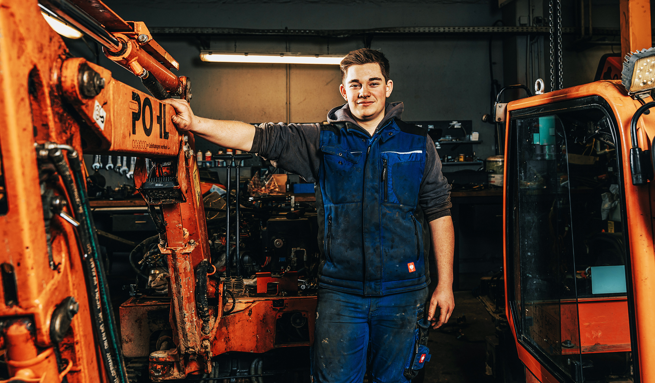 In der Werkstatt lehnt ein POHL-Mechatroniker an einem Baggerarm mit POHL-Schriftzug; um ihn herum Maschinen und Werkzeuge.