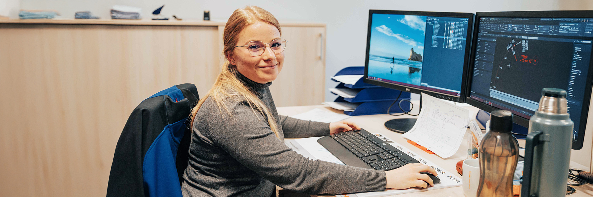 Ausbildung: In der Mitte sitzt eine Frau an einem Schreibtisch und arbeitet an Tastatur und Maus vor zwei Monitoren. Rechts im Bild zeigen die Bildschirme technische Zeichnungen und Listen. Links im Bild liegt eine blaue Jacke über der Stuhllehne. Vorne im Bild stehen Trinkflaschen, Becher und Notizzettel auf dem Tisch. Im Hintergrund steht ein helles Sideboard.