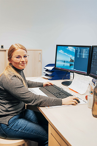 In der Mitte sitzt eine Frau an einem Schreibtisch und arbeitet an Tastatur und Maus vor zwei Monitoren. Rechts im Bild zeigen die Bildschirme technische Zeichnungen und Listen. Links im Bild liegt eine blaue Jacke über der Stuhllehne. Vorne im Bild stehen Trinkflaschen, Becher und Notizzettel auf dem Tisch. Im Hintergrund steht ein helles Sideboard.