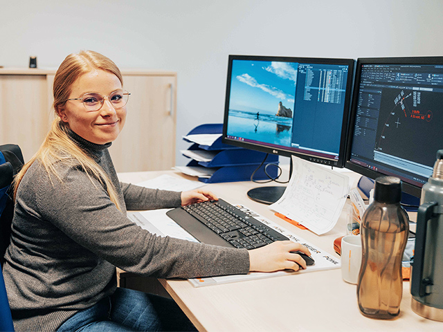 In der Mitte sitzt eine Frau an einem Schreibtisch und arbeitet an Tastatur und Maus vor zwei Monitoren. Rechts im Bild zeigen die Bildschirme technische Zeichnungen und Listen. Links im Bild liegt eine blaue Jacke über der Stuhllehne. Vorne im Bild stehen Trinkflaschen, Becher und Notizzettel auf dem Tisch. Im Hintergrund steht ein helles Sideboard.