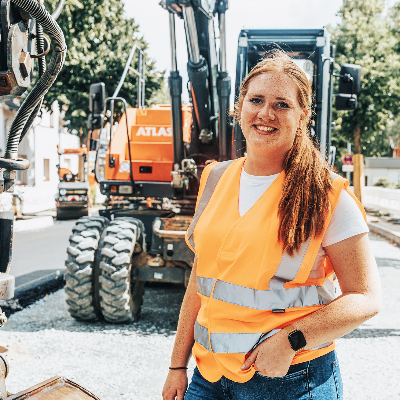 In der Mitte steht eine POHL-Mitarbeiterin mit orange Warnweste auf einer Straßenbaustelle. Links im Vordergrund ist ein graues Hydraulikteil, hinten rechts stehen orange POHL-Baumaschinen; rechts im Hintergrund arbeitet ein weiterer POHL-Mitarbeiter.