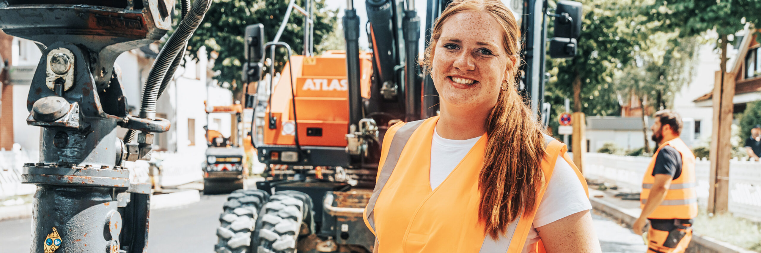 In der Mitte steht eine POHL-Mitarbeiterin mit orange Warnweste auf einer Straßenbaustelle. Links im Vordergrund ist ein graues Hydraulikteil, hinten rechts stehen orange POHL-Baumaschinen; rechts im Hintergrund arbeitet ein weiterer POHL-Mitarbeiter.