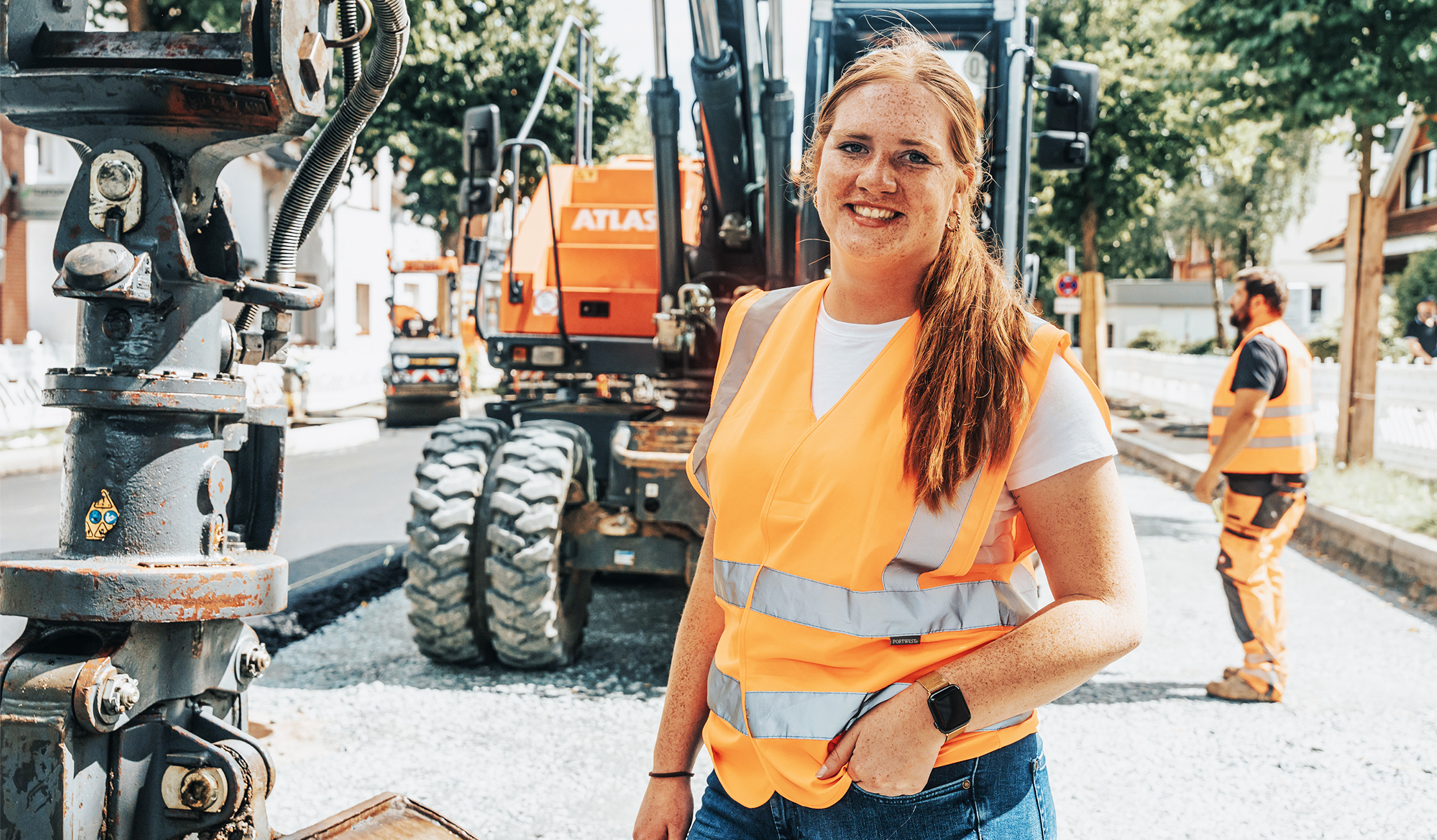 In der Mitte steht eine POHL-Mitarbeiterin mit orange Warnweste auf einer Straßenbaustelle. Links im Vordergrund ist ein graues Hydraulikteil, hinten rechts stehen orange POHL-Baumaschinen; rechts im Hintergrund arbeitet ein weiterer POHL-Mitarbeiter.