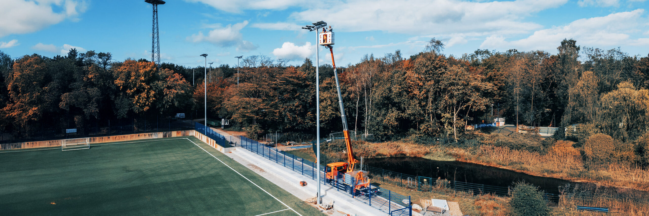 Ein Fußballfeld mit hohen Flutlichtmasten. Ein Pohl-Fahrzeug mit Hubarbeitsbühne steht am Spielfeldrand, zwei Personen arbeiten in großer Höhe an den Scheinwerfern.