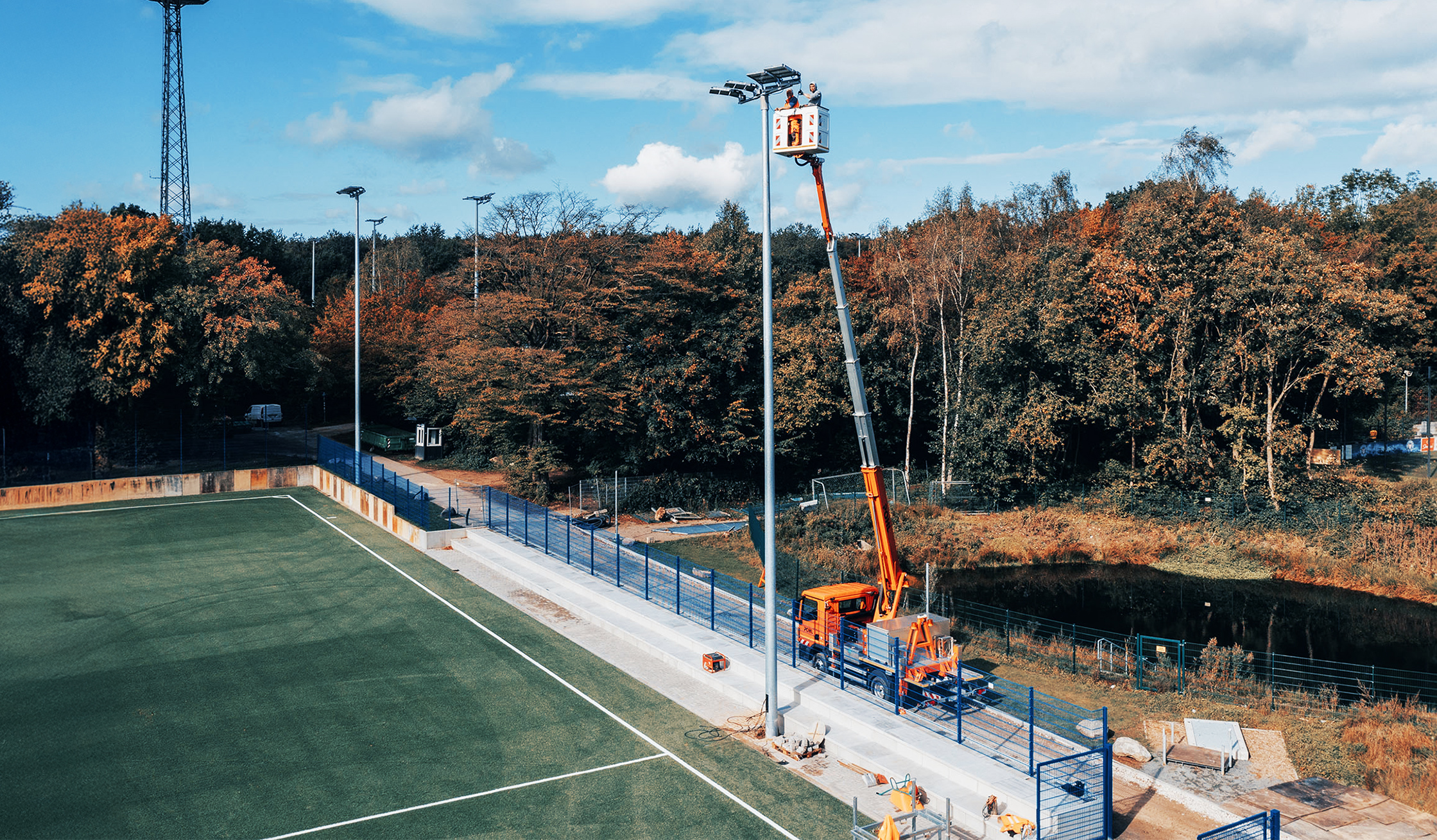 Ein Fußballfeld mit hohen Flutlichtmasten. Ein Pohl-Fahrzeug mit Hubarbeitsbühne steht am Spielfeldrand, zwei Personen arbeiten in großer Höhe an den Scheinwerfern.