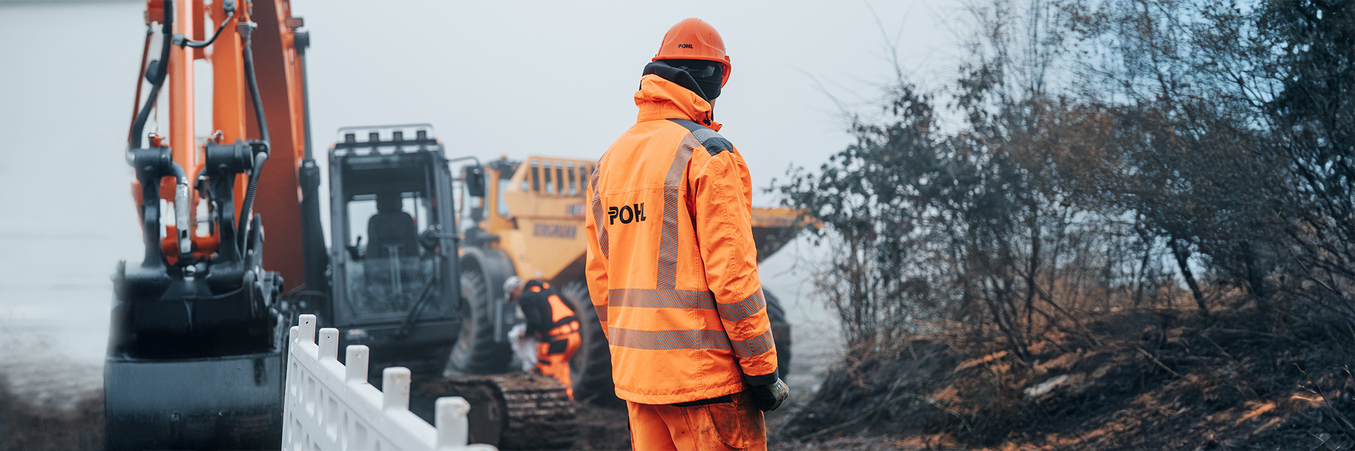 In der Mitte steht ein POHL-Mitarbeiter in orangefarbener Jacke und mit orangefarbenem Helm mit dem Rücken zur Kamera an einer weißen Absperrung. Links im Bild stehen unscharf eine große orangefarbene Baumaschine und weitere Geräte. Rechts im Bild wächst niedriges Buschwerk am Rand. Im Hintergrund arbeiten weitere Personen zwischen Maschinen auf hellem Boden.