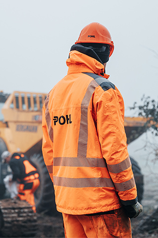 In der Mitte steht ein POHL-Mitarbeiter in orangefarbener Jacke und mit orangefarbenem Helm mit dem Rücken zur Kamera an einer weißen Absperrung. Links im Bild stehen unscharf eine große orangefarbene Baumaschine und weitere Geräte. Rechts im Bild wächst niedriges Buschwerk am Rand. Im Hintergrund arbeiten weitere Personen zwischen Maschinen auf hellem Boden.