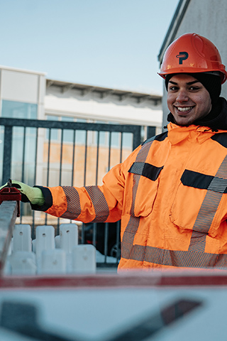 In der Mitte steht ein POHL-Mitarbeiter in orangefarbener Jacke und Helm und blickt zur Kamera; links vorne verläuft eine weiße Absperrung vor einem Gebäude mit großen Fenstern, rechts am Rand ist eine graue Wand.