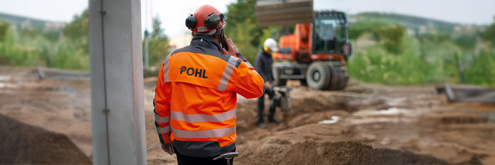 In der Mitte steht ein POHL-Mitarbeiter mit orangefarbener Jacke und Helm und telefoniert; rechts im Hintergrund ist unscharf ein orangefarbener POHL-Bagger auf einer Erdfläche zu sehen.