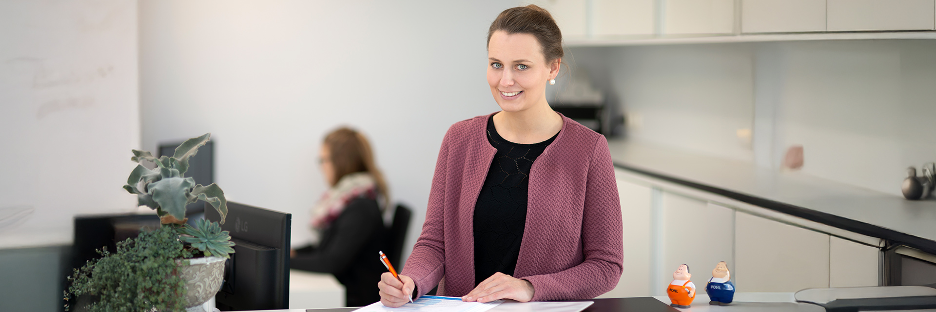 In der Mitte steht eine Frau an einem Tresen und schreibt auf ein Blatt; links im Bild steht ein Topf mit Pflanzen, rechts auf dem Tresen stehen zwei kleine Figuren mit POHL-Aufdruck, im Hintergrund arbeitet eine Person am Schreibtisch.