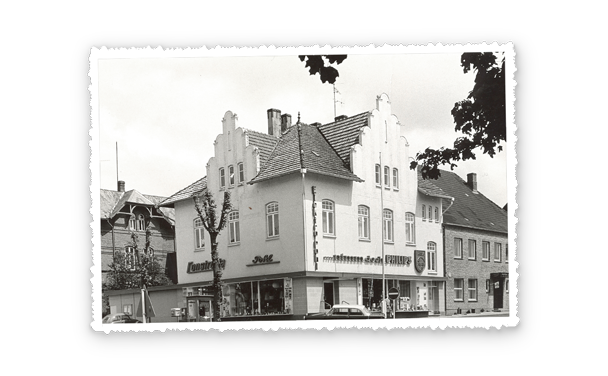 Schwarzweißaufnahme eines Geschäftsgebäudes an einer Straßenecke mit markanter Giebelfassade, im Erdgeschoss liegen Schaufenster.